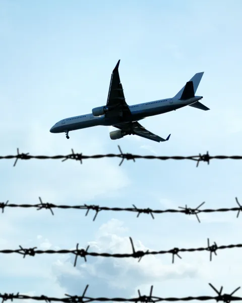 A shot looking up at a plane flying in a blue sky over a barbed wire fence in the foreground.