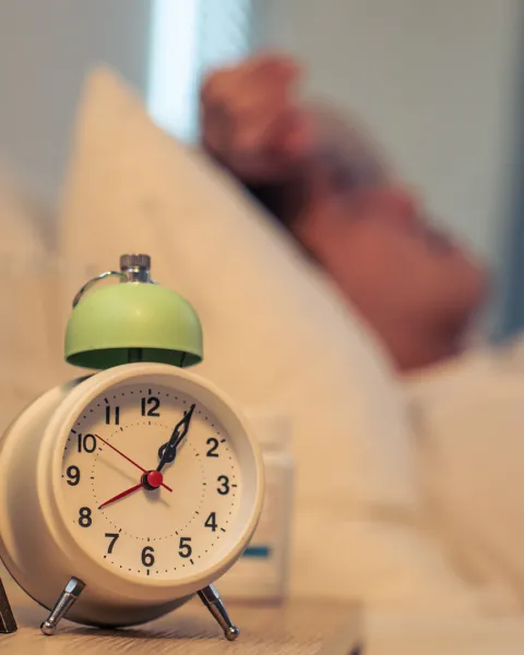 Close-up of an alarm clock on nightstand with sick man laying in bed in background.