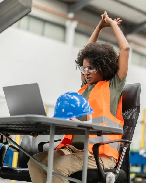 Tired female industrial engineer stretching arms at desk in factory.