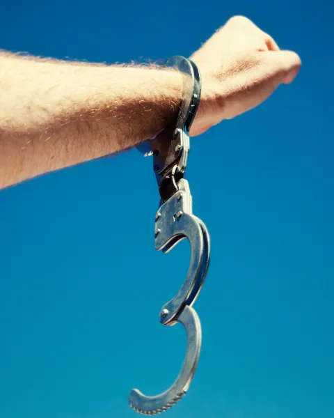 A man's hand, holding up a clenched fist in a salute, and wearing a single, open handcuff, against clear blue sky