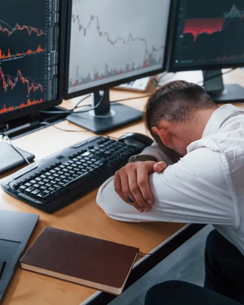 Tired businessman head down on his desk with multiple screens on it which display stock information.