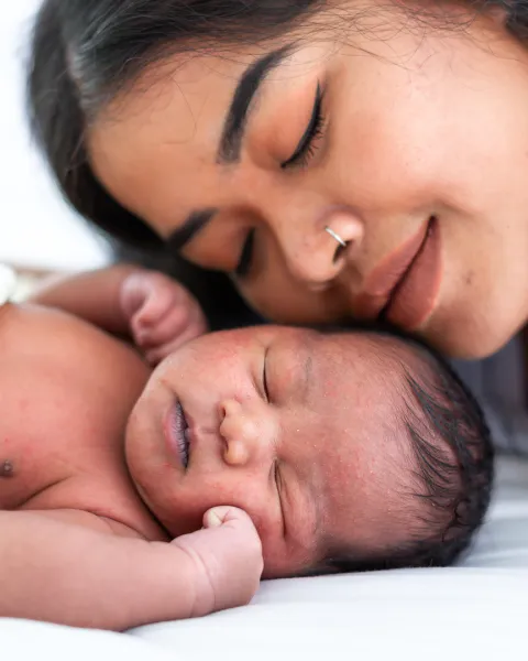 Ethnic mother hugging and kissing her newborn baby on white bed. 