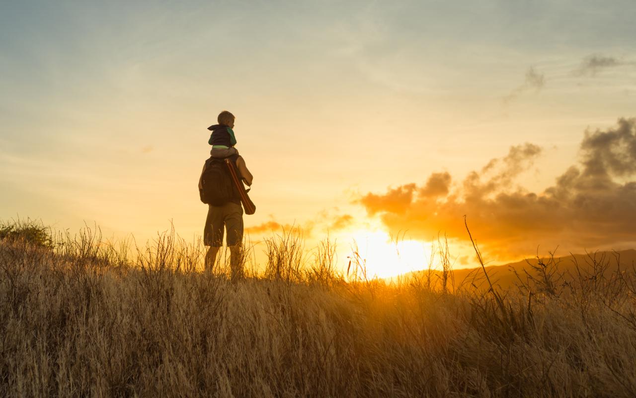 Father with son on his shoulders, hiking through a field at sunrise.
