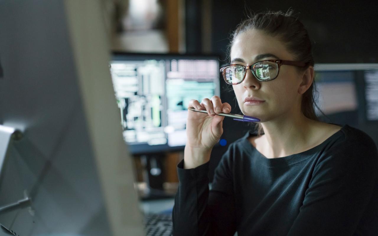 Image of a young woman’s face as she contemplates one of the many computer monitors that surround her.