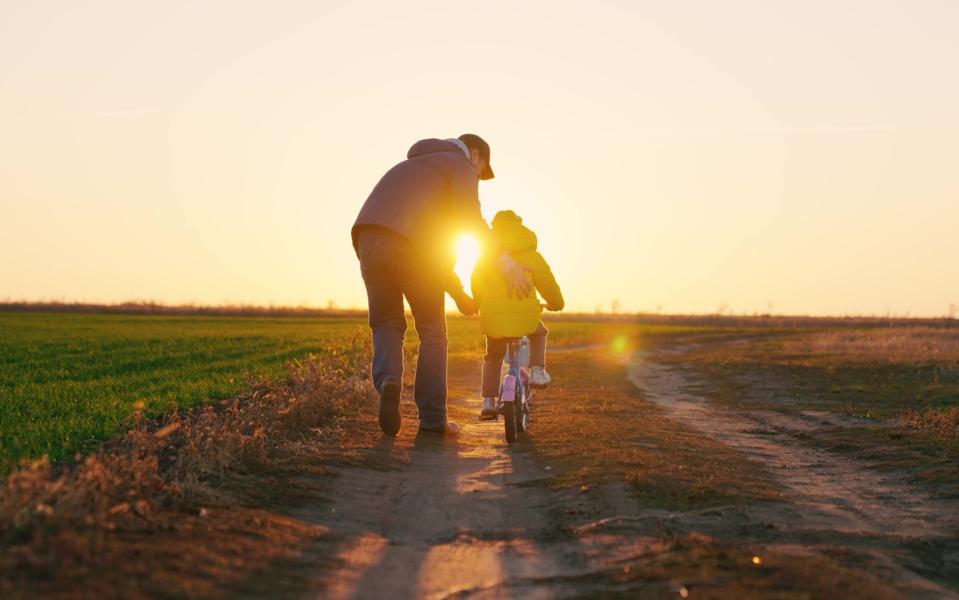 A father teaches his daughter to ride a bike in the park at sunset.