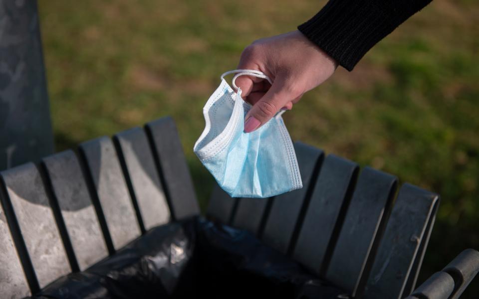 A close-up of a hand throwing a face mask into a garbage can.