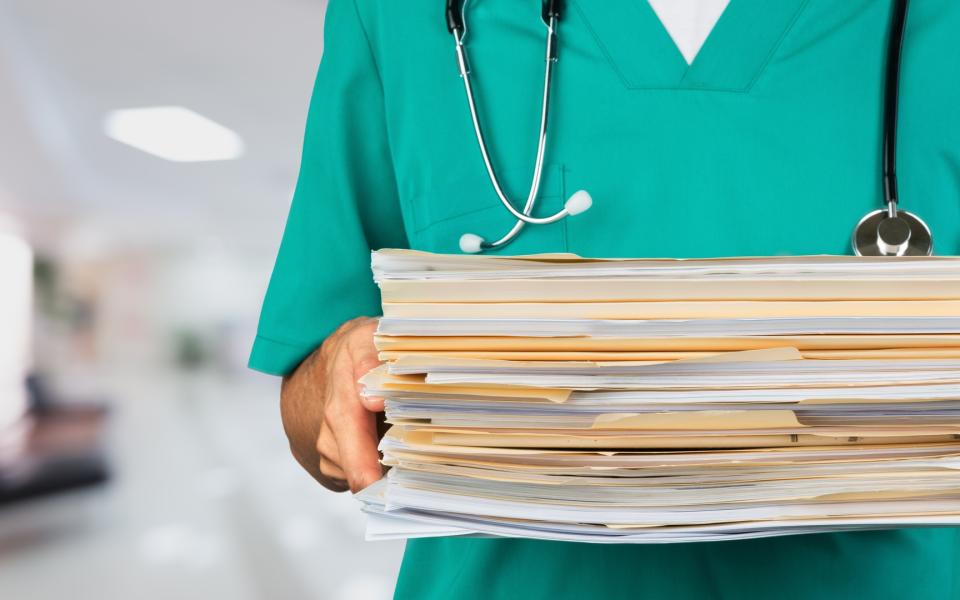 A close-up of a doctor in scrubs holding stacks of files/paperwork.