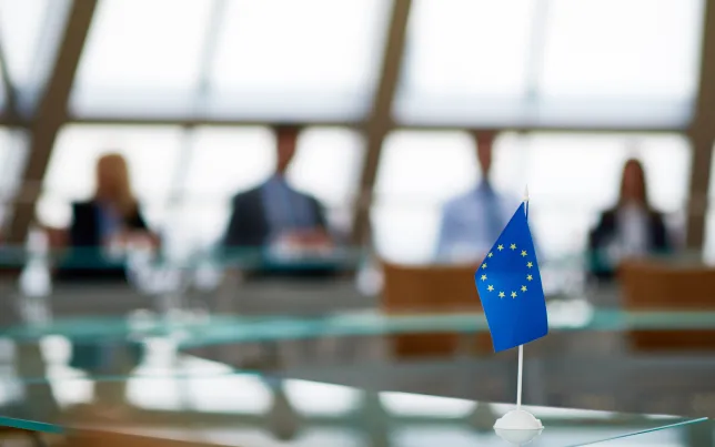 Soft-focus view of a boardroom and a small EU flag sitting on the desk in the foreground.