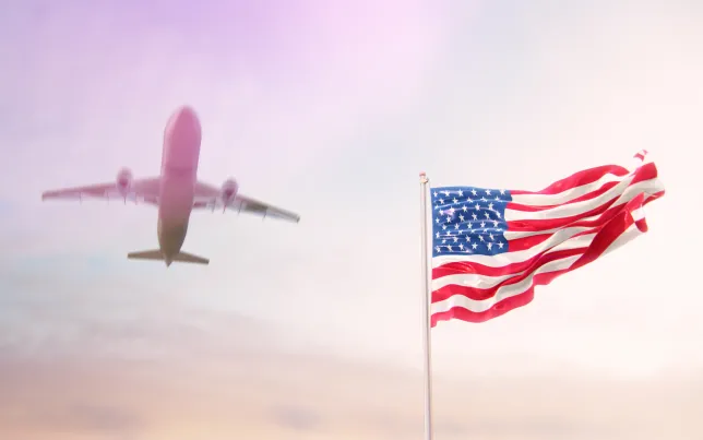 American flag blowing on a pole with an airplane flying above it in the sky.