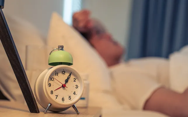 Close-up of an alarm clock on nightstand with sick man laying in bed in background.