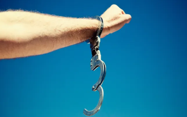 A man's hand, holding up a clenched fist in a salute, and wearing a single, open handcuff, against clear blue sky
