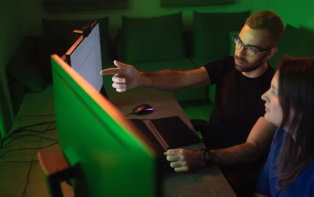 A pair of software developers look at multiple computer monitors gesturing at, and discussing what they see.