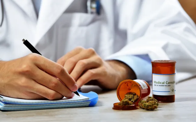 Close-up of a doctor writing on a prescription pad with a prescription bottle and medical cannabis on the table.