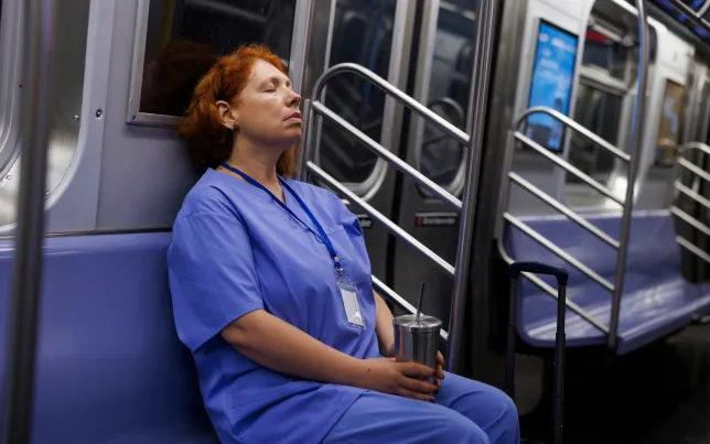 Resting her eyes during the evening commute, a middle-aged nurse in scrubs sits on a subway train.
