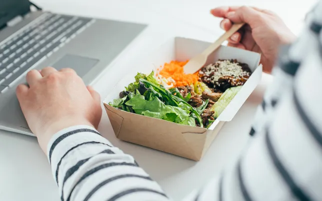 Close-up of a woman's hands eating a boxed lunch in front of her laptop.