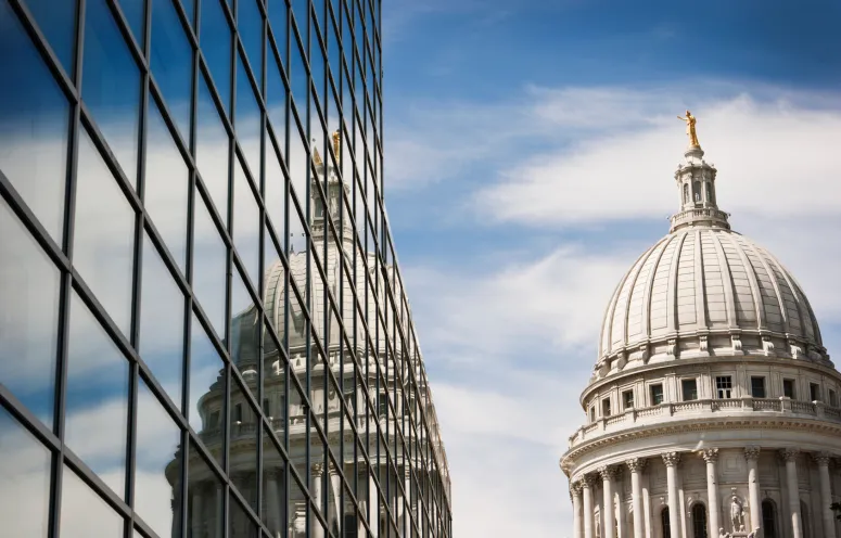 Horizontal view of the Wisconsin State Capitol Dome reflecting into the windows of a steel and glass office building on a sunny day.